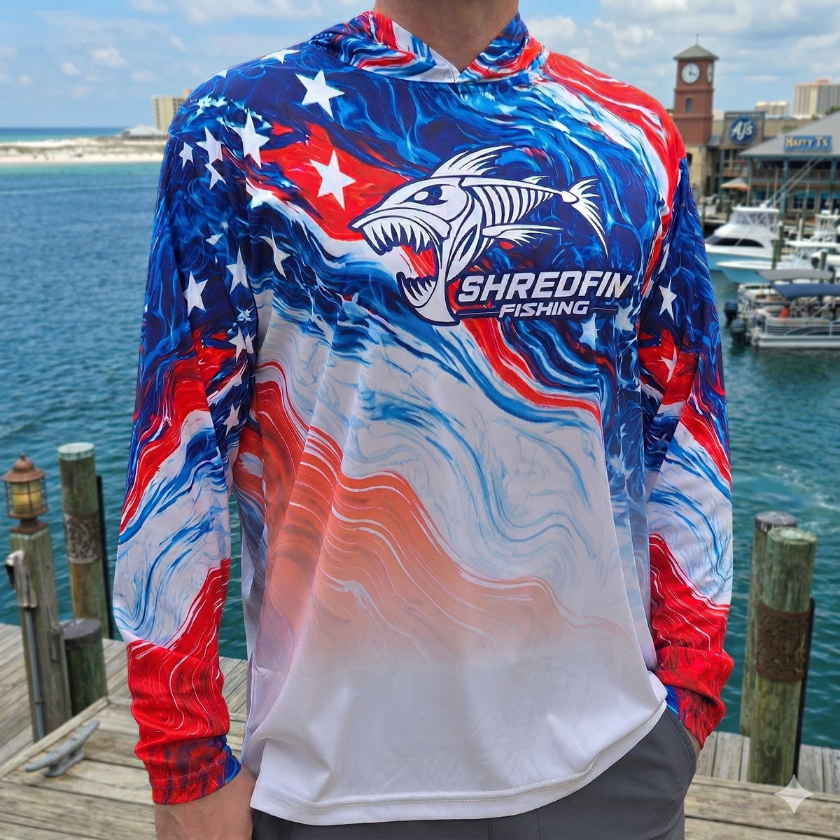 Man wearing a ShredFin patriotic red, white, and blue hooded performance fishing shirt with UPF 50+ sun protection at a marina.