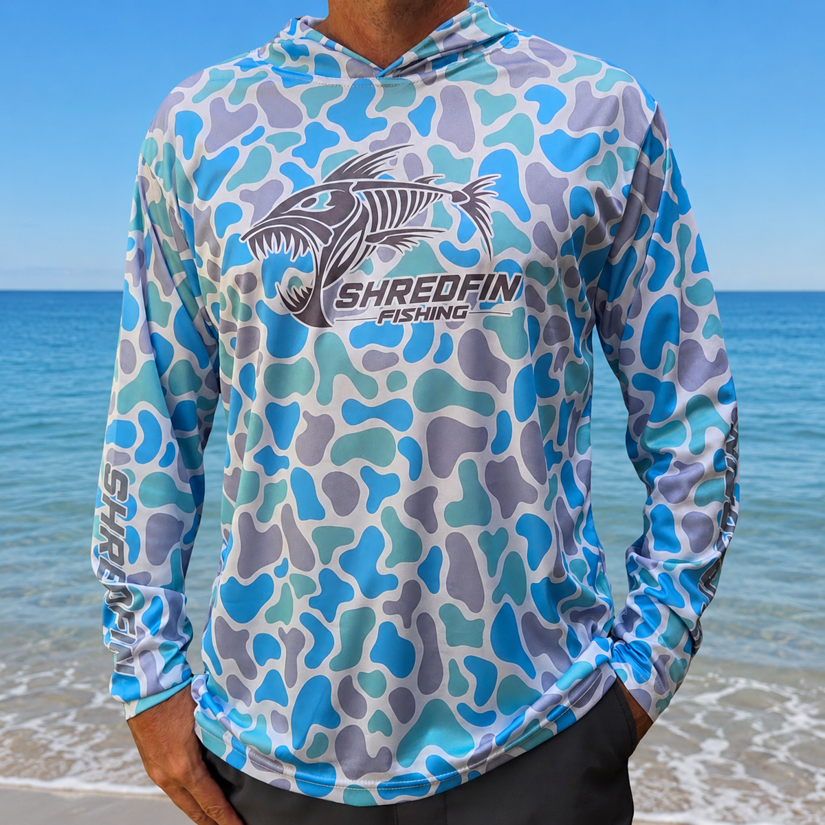 Man wearing a ShredFin Retro Camo Sea Glass hooded performance fishing shirt on a beach with an ocean backdrop.