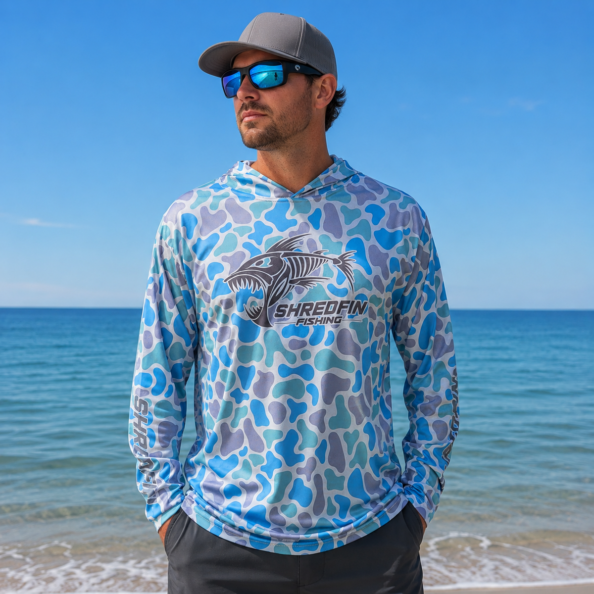 Man wearing a ShredFin Retro Camo Sea Glass hooded performance fishing shirt on a beach with blue mirrored sunglasses and an ocean backdrop.