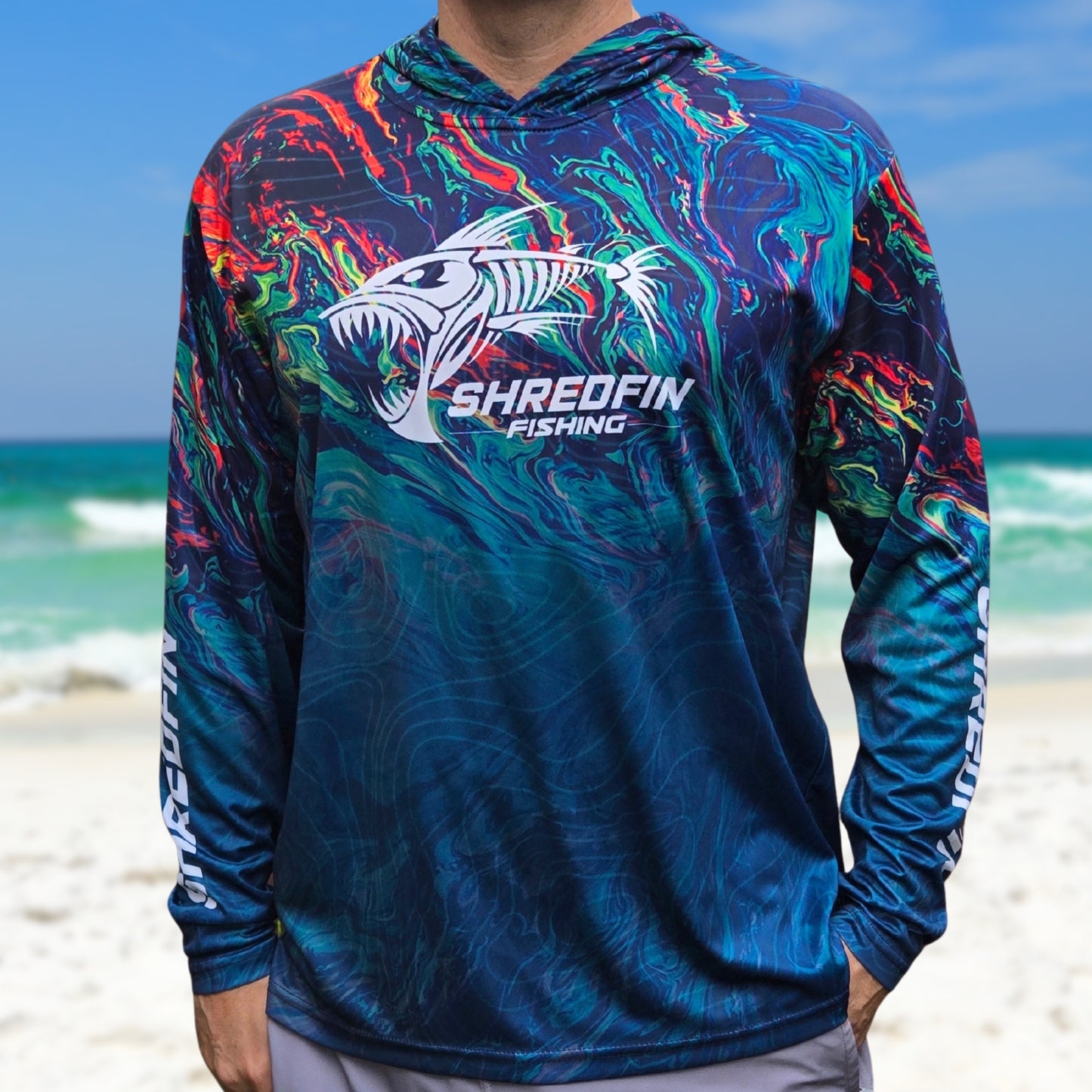 Man wearing a ShredFin StormSurge Electric Swirl hooded performance fishing shirt on a beach featuring a blue, teal, and orange ocean-inspired pattern.